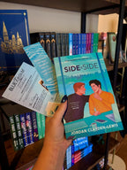 A hand with blue glitter nail polis holds the book 'Side by Side' by Jordan Clayden-Lewis, a bookmark and postcard in front of a bookshelf. The cover features an illustrated scene of two men sitting at a desk with a laptop and coffee mug, with a city skyline in the background.
