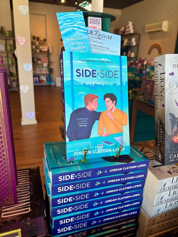 A stack of paperback books titled Book cover for 'Side by Side' by Jordan Clayden-Lewis displayed on a store shelf, with a matching blue promotional bookmark and postcard leaning behind the top copy. The books feature an illustrated scene of two men sitting at a desk with a laptop and coffee mug, with a city skyline in the background.