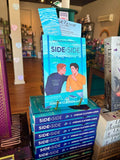 A stack of paperback books titled Book cover for 'Side by Side' by Jordan Clayden-Lewis displayed on a store shelf, with a matching blue promotional bookmark and postcard leaning behind the top copy. The books feature an illustrated scene of two men sitting at a desk with a laptop and coffee mug, with a city skyline in the background.