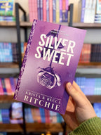 A hand holding a paperback copy of 'Silver Sweet' by Krista & Becca Ritchie with a purple background and rose design, and matching sprayed edges with purple roses.