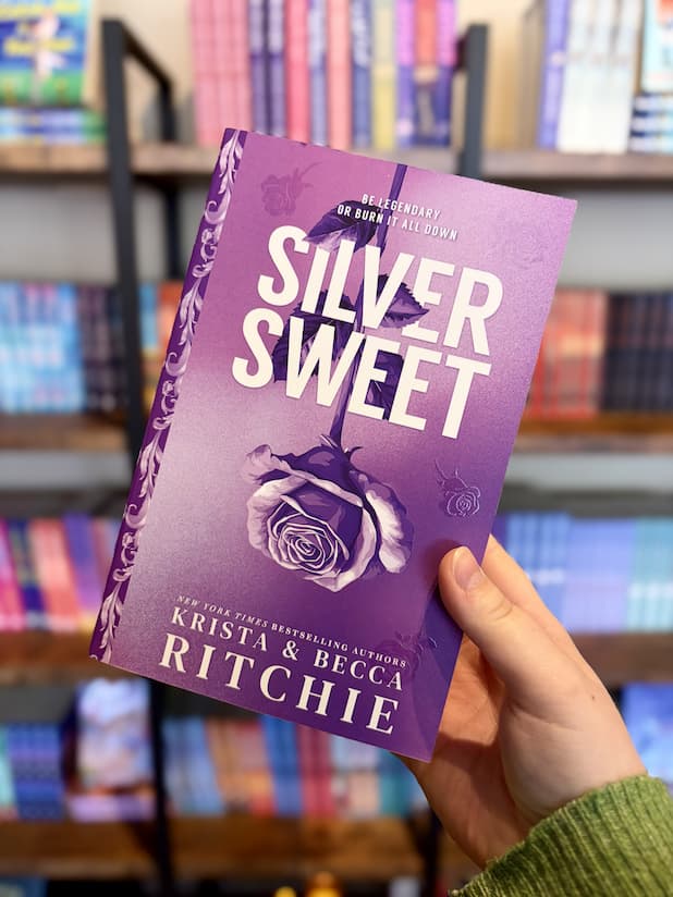 A hand holding a paperback copy of 'Silver Sweet' by Krista & Becca Ritchie with a purple background and rose design, and matching sprayed edges with purple roses.