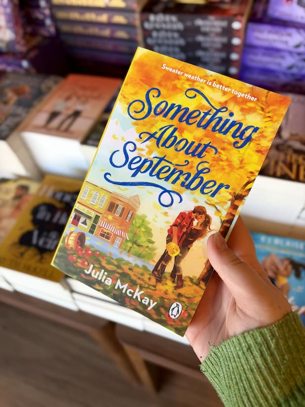 A hand holding a copy of 'Something About September' by Julia McKay with a couple kissing under a tree, surrounded by autumn leaves.