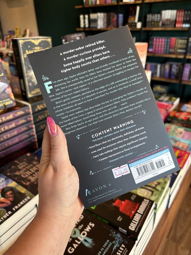A hand with pink nail polish holding the back of a copy of 'Songbird in the Gallows' by Alta Hensley, featuring a black background and blue and white text.