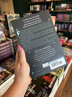 A hand with pink nail polish holding the back of a copy of 'Songbird in the Gallows' by Alta Hensley, featuring a black background and blue and white text.