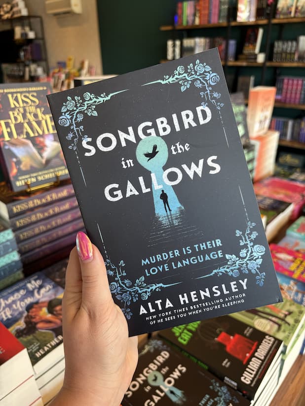 A hand with pink nail polish holding a copy of 'Songbird in the Gallows' by Alta Hensley, featuring a human silhouette inside an illuminates large keyhole with a floral border design, and white text.