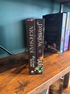 Sorrowsong University Books One and Two standing upright on a wooden shelf: 'Nightshade' by Autumn Woods on the left and 'Daybreak' by Autumn Woods on the right, with other books blurred in the background against a teal wall.