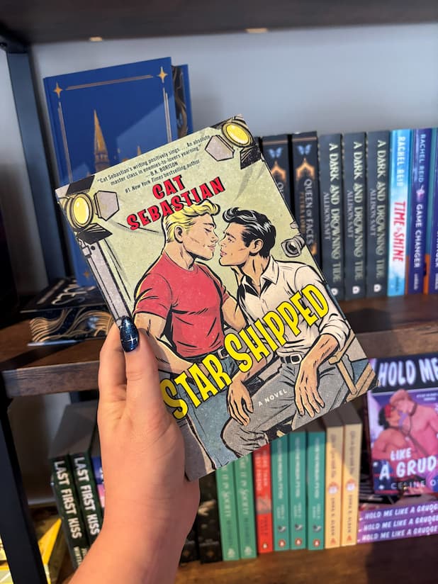 A hand holds the book cover for the novel 'Star Shipped' by Cat Sebastian, featuring a retro comic-style illustration of two men on a film set about to kiss with large yellow title text across the bottom in front of a blurry wooden bookshelf.