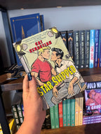 A hand holds the book cover for the novel 'Star Shipped' by Cat Sebastian, featuring a retro comic-style illustration of two men on a film set about to kiss with large yellow title text across the bottom in front of a blurry wooden bookshelf.
