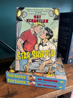 The book cover for the novel 'Star Shipped' by Cat Sebastian sits on a gold stand on top of a stack of the same book on a wooden bookshelf, featuring a retro comic-style illustration of two men on a film set about to kiss with large yellow title text across the bottom.
