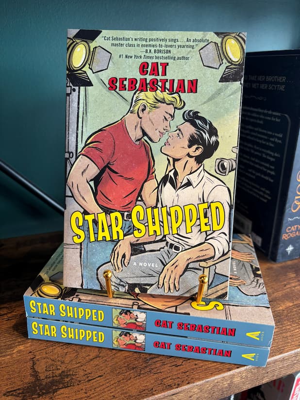 The book cover for the novel 'Star Shipped' by Cat Sebastian sits on a gold stand on top of a stack of the same book on a wooden bookshelf, featuring a retro comic-style illustration of two men on a film set about to kiss with large yellow title text across the bottom.