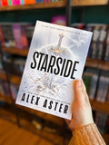 A hand holding a copy of the book 'Starside' by Alex Aster in a bookstore, with bookshelves blurred in the background.