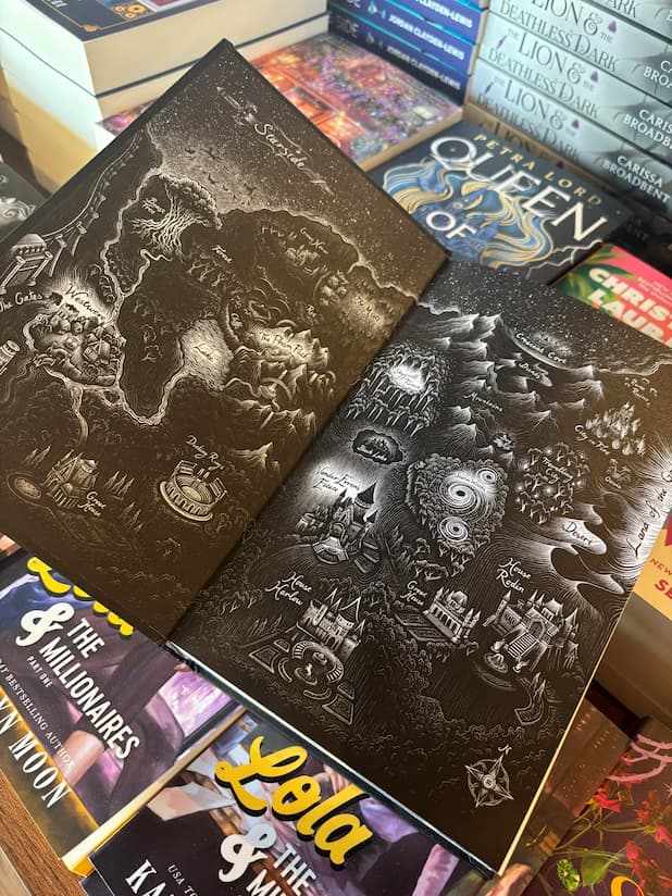 A hand holds open the special edition version of the book 'Starside' by Alex Aster, featuring a black and white illustrated map sitting on a table full of other books blurry in the background.