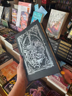 A holds the special edition version of the book 'Starside' by Alex Aster with the dust jacket removed, featuring a silver foiled image of a woman holding a sword. The book is held in front of a table full of other books blurry in the background. 