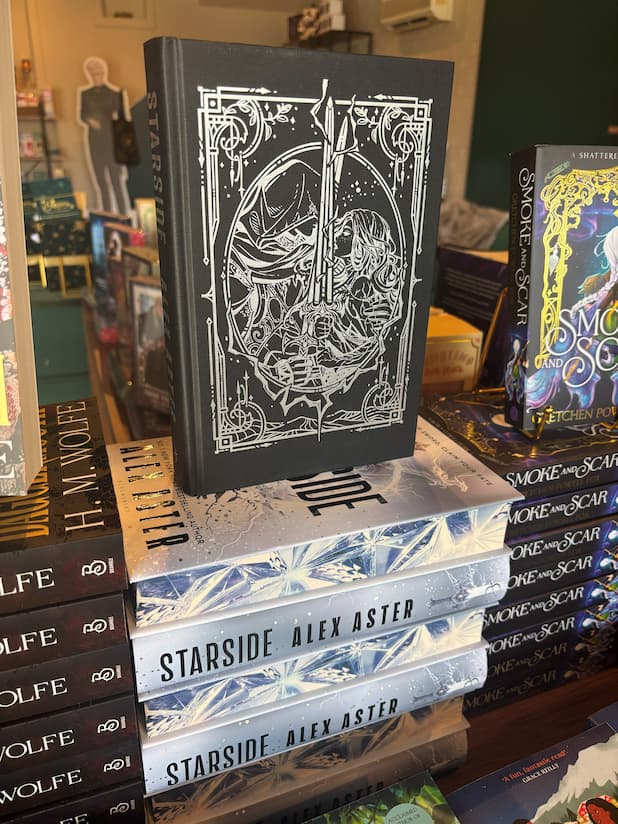 A stack of the special edition version of the book 'Starside' by Alex Aster, featuring a large silver sword piercing icy shards with lightning effects, and crystalline digital printed edges, alternating spine and digitally printed edges, the top book has the dust jacket removed and has a silver foiled image of a woman holding a sword.