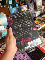A hand holds the back cover of Steel Princess by Rina Kent, Book Two in the Royal Elite Series. White and purple font are used for the3 blurb on top of a black floral background with pops of pink and purple flowers. The book is held in front of a table full of other books blurry in the background. 