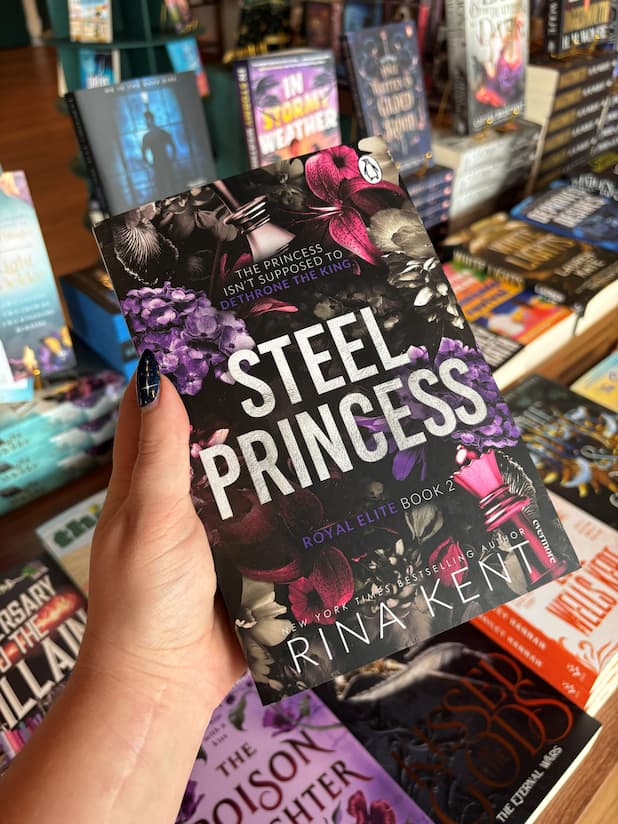 A hand holds the book 'Steel Princess' by Rina Kent in a bookstore aisle, surrounded by other books on shelves.
