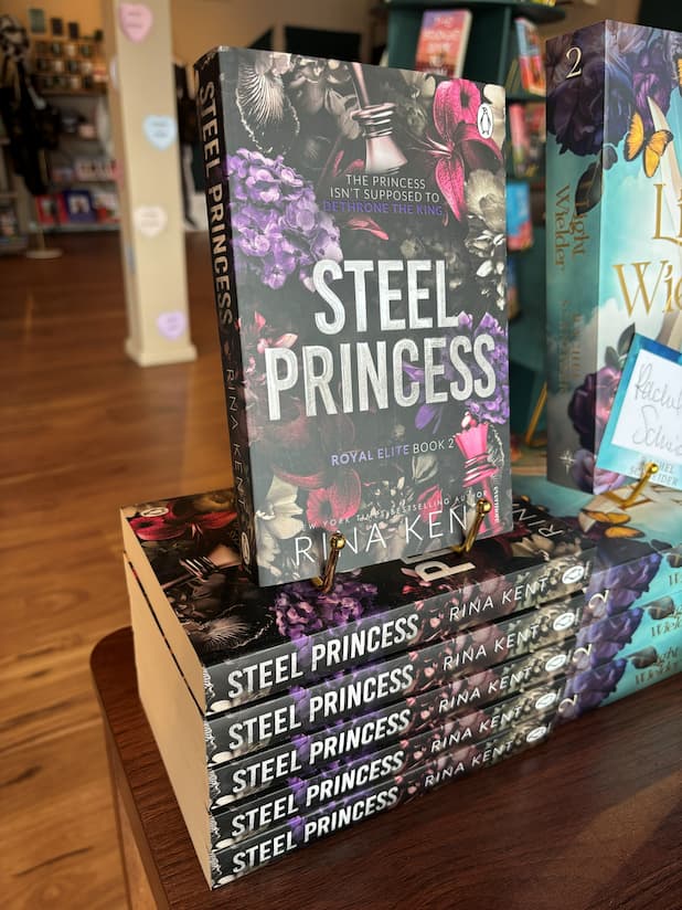 A stack of the book Steel Princess by Rina Kent, Book Two in the Royal Elite Series. Metallic font overlaps a maroon, black and purple floral pattern with a king and queen chess piece in opposite corners. The books sit on a wooden table in a bookstore with other books blurry in the background.
