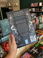 A hand with pink nail polish holding a copy of 'Styx the River' by Nikita Gill, featuring two blue female figures standing in stylized a black background with silver seaweed, fish, and text.