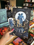 A hand with pink nail polish holding a copy of 'Styx the River' by Nikita Gill, featuring two blue female figures standing in stylized waves against a black background with white decorative elements and stars, and a matching bookmark.