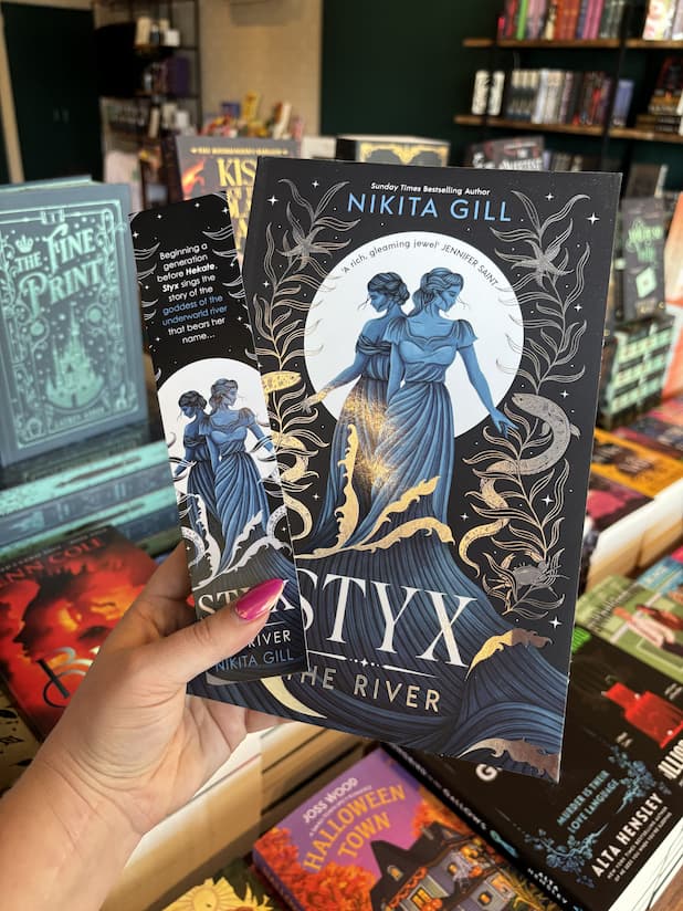 A hand with pink nail polish holding a copy of 'Styx the River' by Nikita Gill, featuring two blue female figures standing in stylized waves against a black background with white decorative elements and stars, and a matching bookmark.