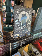 A stack of the book 'Styx the River' by Nikita Gill, featuring two blue female figures standing in stylized waves against a black background with white decorative elements and stars.