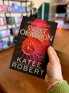 A hand holding a paperback copy of the book 'Sweet Obsession' by Katee Robert. It is featuring a glowing pink and red spiral shell on a dark background with white and green title text.