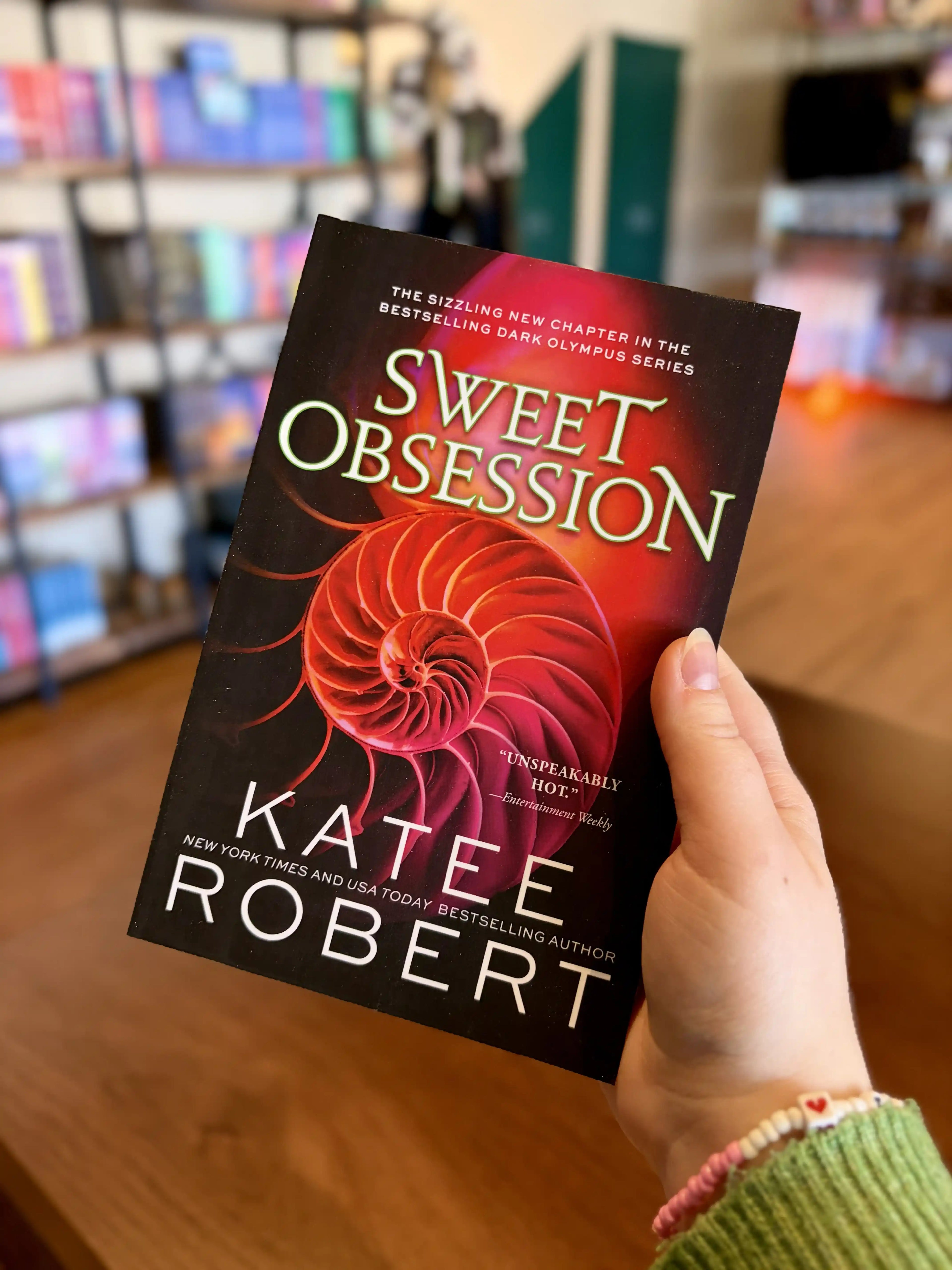 A hand holding a paperback copy of the book 'Sweet Obsession' by Katee Robert. It is featuring a glowing pink and red spiral shell on a dark background with white and green title text.
