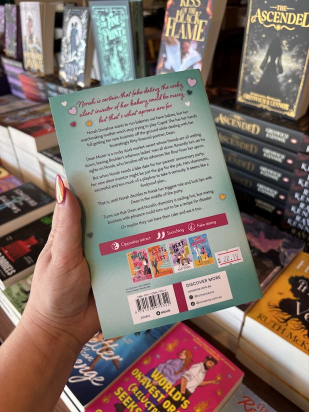 A hand with pink nail polish holding the back of a copy of 'Take a Number' by Amy Daws, featuring a pastel teal background with pink and black text.