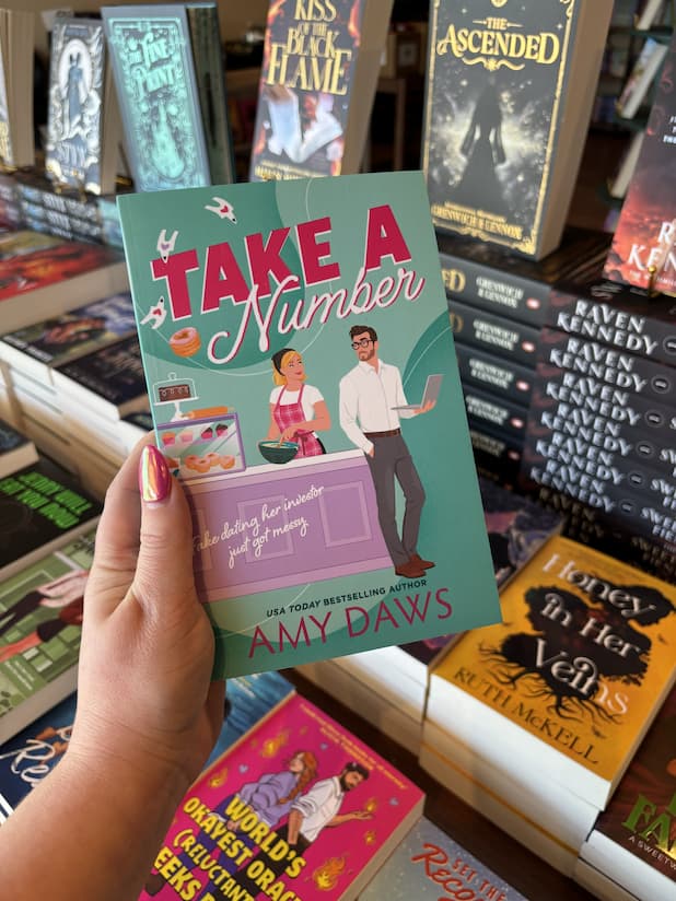 A hand with pink nail polish holding a copy of 'Take a Number' by Amy Daws, featuring a man and woman at a bakery counter.