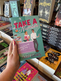 A hand with pink nail polish holding a copy of 'Take a Number' by Amy Daws, featuring a man and woman at a bakery counter.
