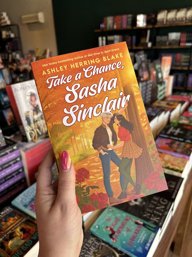 A hand with pink nail polish holding a copy of 'Take a Chance, Sasha Sinclair' with a couple of women in a forest surrounded by roses. One has her back to a tree and looking into the eyes of the other who is leaning against the tree with one arm.