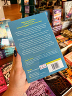 A hand with black glitter nail polish holds the back cover of a book titled 'Take a Hike' by Maggie Grant in a bookstore. The book cover is a light blue color with white text and a barcode, with other books visible in the blurred background.