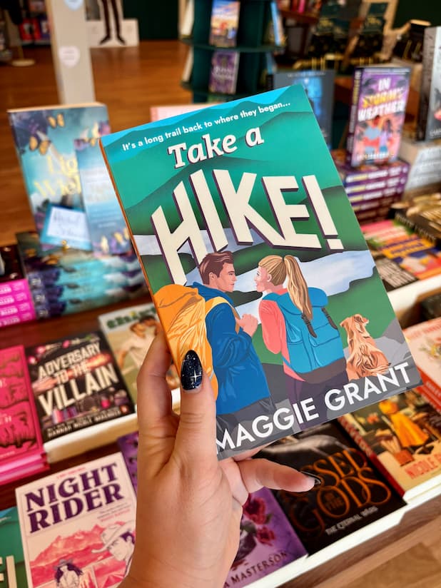 A hand holds the book 'Take a Hike!' by Maggie Grant in a bookstore, with other book covers visible in the background.
