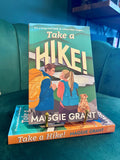 A book titled "Take a Hike!" by Maggie Grant is displayed upright on a teal couch, supported by two small gold bookends. The cover shows an illustrated couple and dog with hiking gear, and a second copy of the book lies underneath.