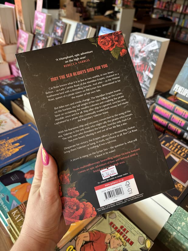 A hand with pink nail polish holds the back of the book 'Tempest' by Victoria Aveyard, featuring a dark background with illustrated roses and white and red text.
