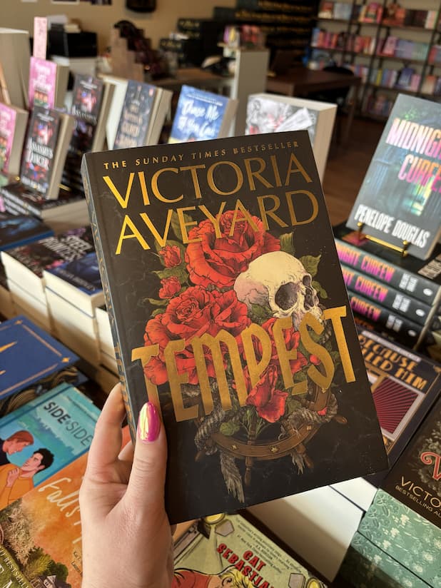 A hand holds a copy of the book 'Tempest' by Victoria Aveyard in a book store. The book cover is black with gold lettering and features red roses and a skull illustration.