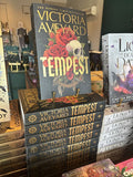 A stack of copies of the book Tempest by Victoria Aveyard displayed on a store shelf, with the front copy standing upright and showing a dark cover with red roses, a skull, and large gold title text.