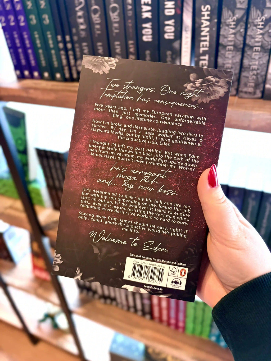 A hand with red nail polish holds a paerback copy of the book 'Tempted by Eden' by Jade May in front of a blurred bookshelf with the back cover facing the camera. The back cover is dark red with a white font and flowers.