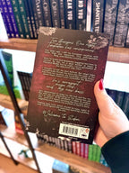 A hand with red nail polish holds a paerback copy of the book 'Tempted by Eden' by Jade May in front of a blurred bookshelf with the back cover facing the camera. The back cover is dark red with a white font and flowers.