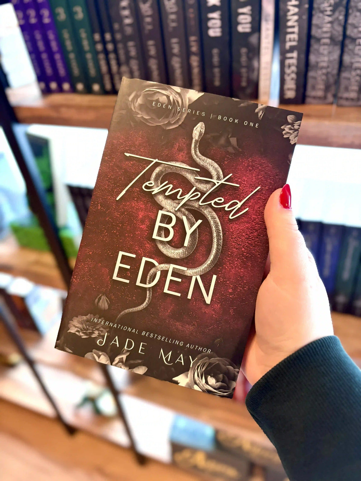 A hand with red nail polish holds a paperback copy of the book 'Tempted by Eden' by Jade May in front of a blurred bookshelf. The cover is dark red with a grey snake in the centre and grey roses on the top and bottom of the cover.