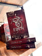 A stack of three copies of the book 'Tempted by Eden' by Jade May, displayed on a wooden bookshelf. The cover is dark red with a grey snake in the centre and grey roses on the top and bottom of the cover.