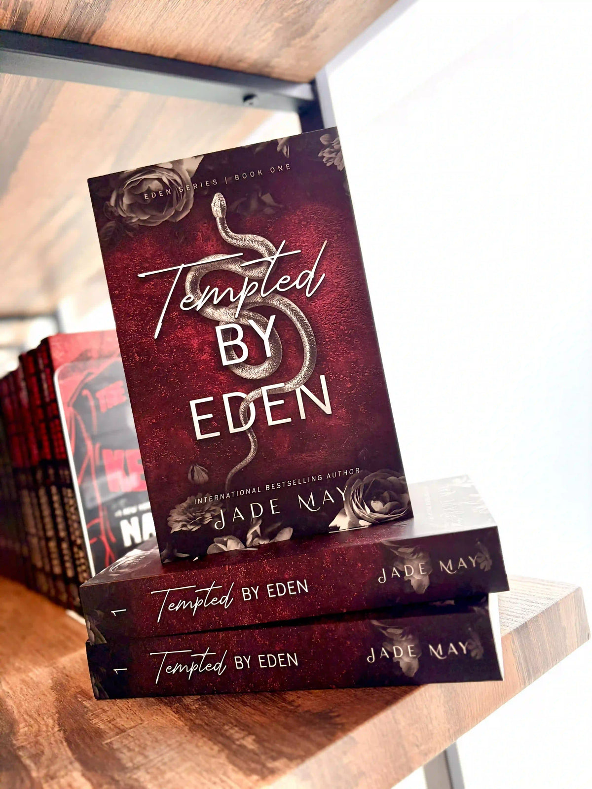 A stack of three copies of the book 'Tempted by Eden' by Jade May, displayed on a wooden bookshelf. The cover is dark red with a grey snake in the centre and grey roses on the top and bottom of the cover.