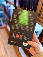 A hand holding the back of a paperback copy of the book 'Tender Cruelty' by Katee Robert. The book cover is black with a gold lion's head knocker illustration and white and pink text, with a blurred bookshelf background.