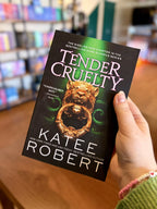 A hand holding a paperback copy of the book 'Tender Cruelty' by Katee Robert. The book cover is black with a gold lion's head knocker illustration and white and pink text, with a blurred bookshelf background.