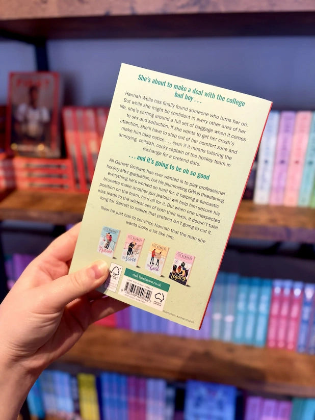 A hand holding the back cover of a paperback book of 'The Deal' by Elle Kennedy. The book cover is cream-colored with black text and small illustrated thumbnails along the bottom.