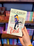 A hand holds the book 'The Deal' by Elle Kennedy in front of a blurred bookshelf background.