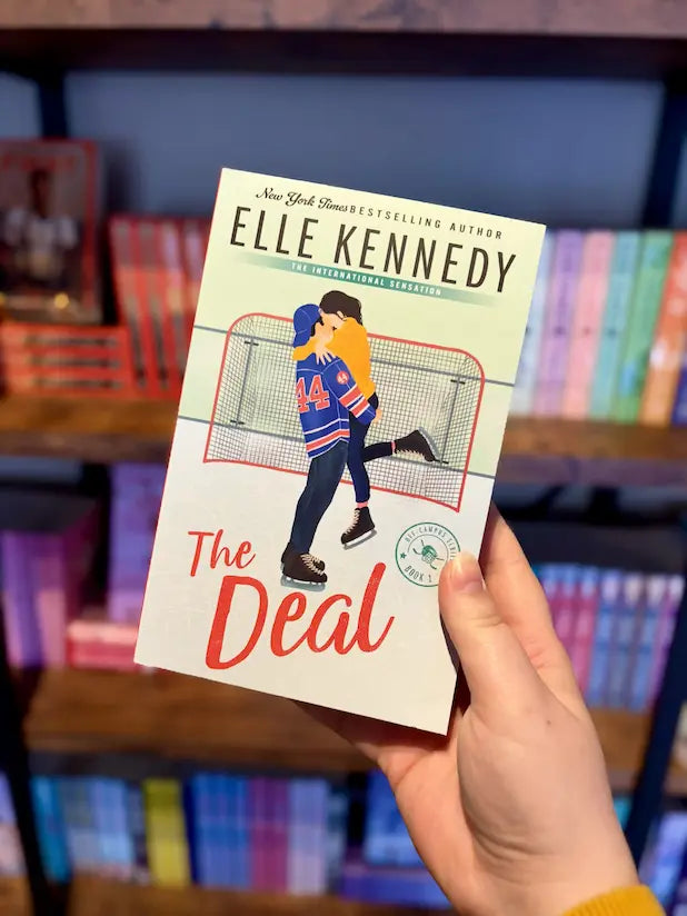 A hand holds the book 'The Deal' by Elle Kennedy in front of a blurred bookshelf background.