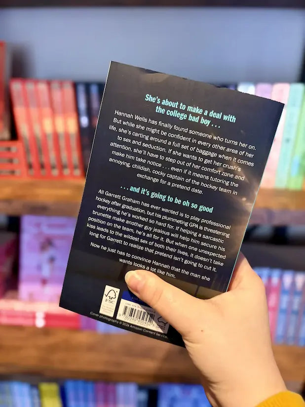A hand holding the back cover of the book 'The Deal' Elle Kennedy in front of a blurred bookshelf background. The book cover is dark with white text and a barcode at the bottom.