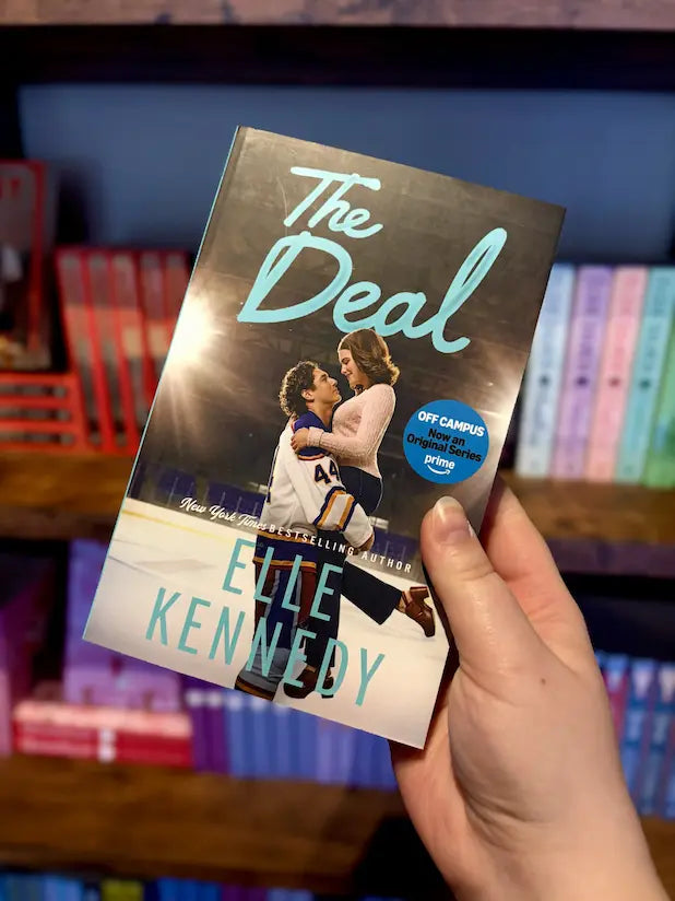 A hand holds the paperback book 'The Deal' by Elle Kennedy in front of a blurred bookshelf background. The cover features a dark hockey-themed design with a couple embracing on ice and a blue circular 'Off-Campus' badge.