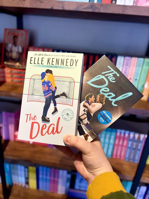 A hand holding two copies of the book 'The Deal' by Elle Kennedy in front of a blurred bookshelf background.